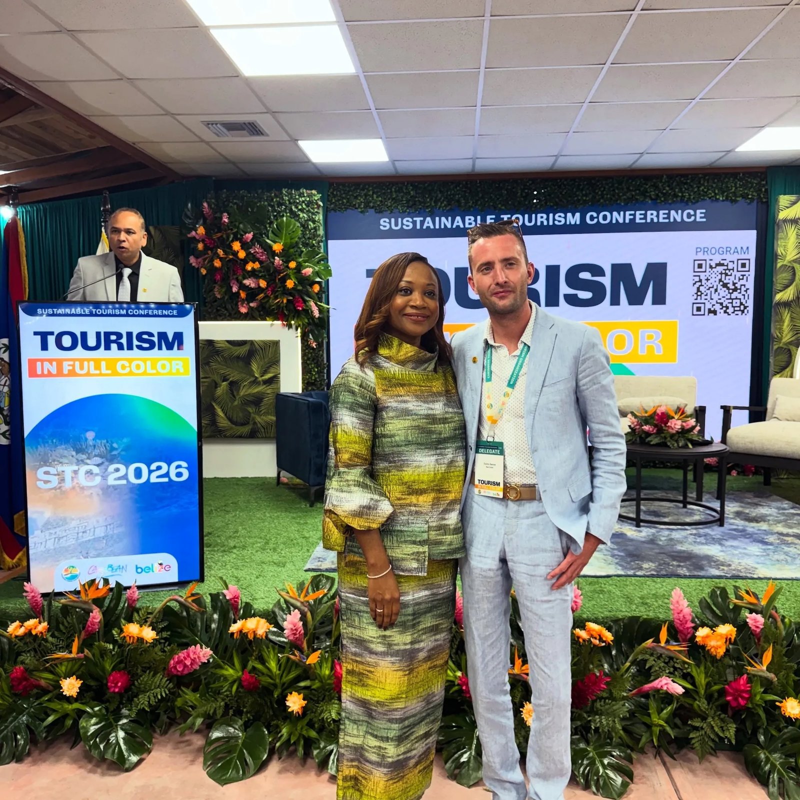 Two conference attendees pose in front of a stage with a blue banner reading 'SUSTAINABLE TOURISM CONFERENCE' and colorful flower arrangements nearby.